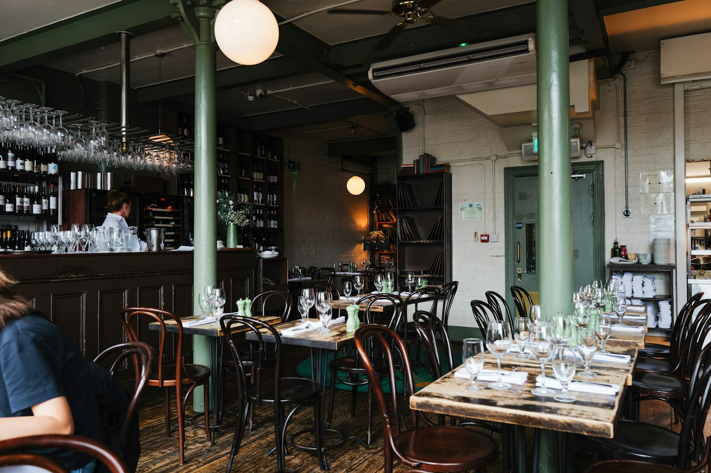 Interior del comedor con mesas de madera dispuestas con copas, sillas curvadas, columnas verdes y barra de fondo