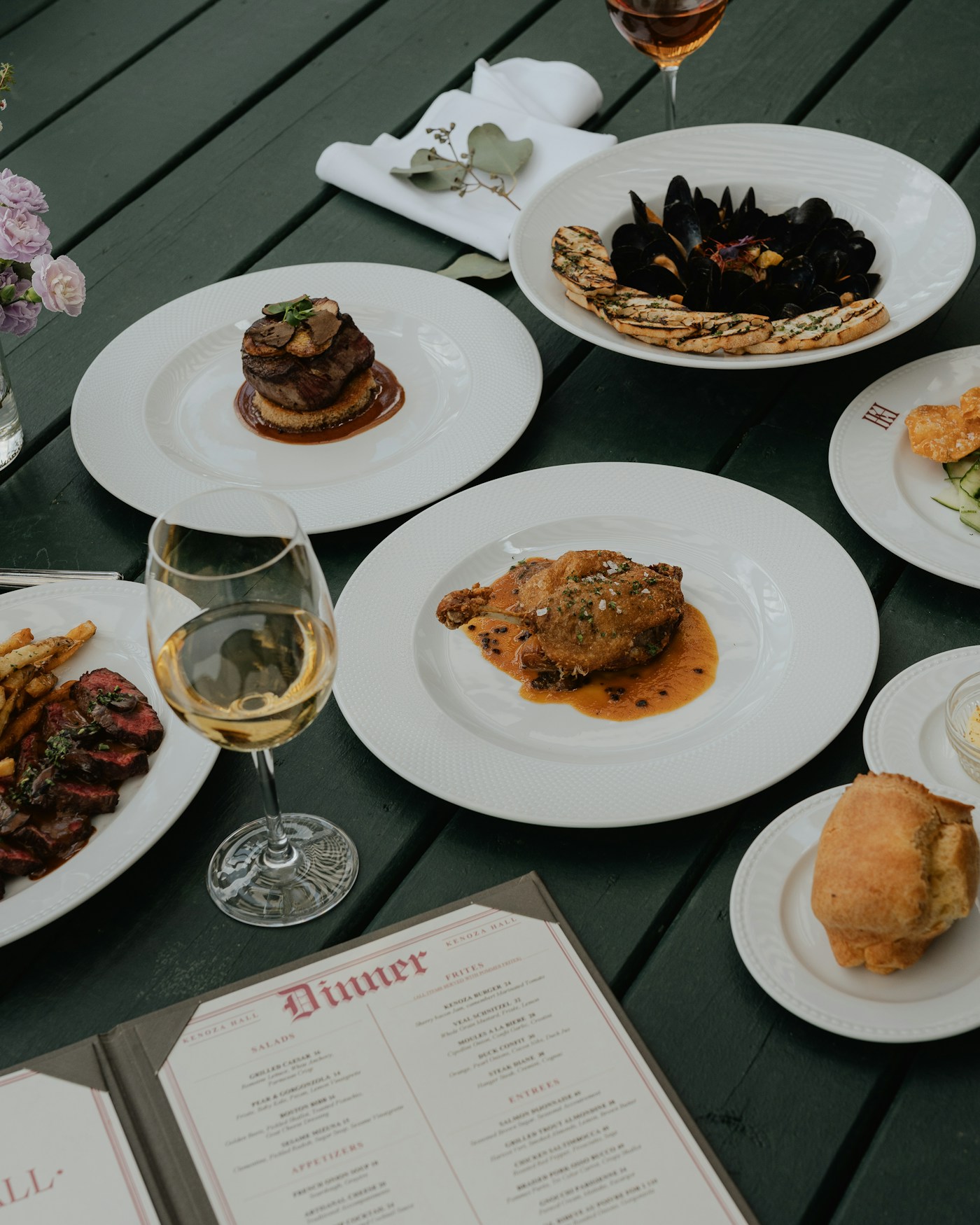 Mesa de madera con variedad de platos degustación, copa de vino y carta abierta